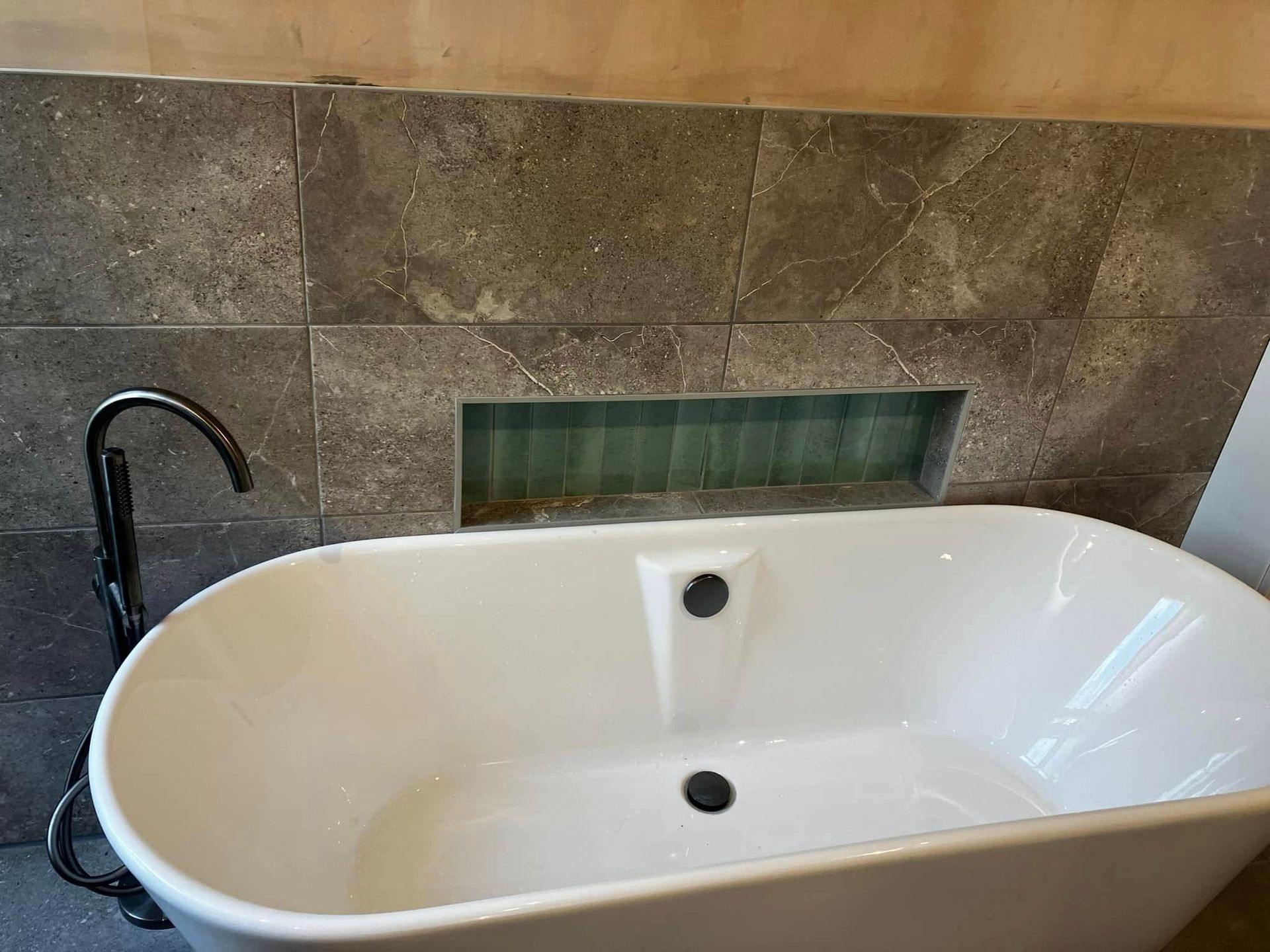 White bathtub with black faucet against gray tiled wall with recessed shelf.