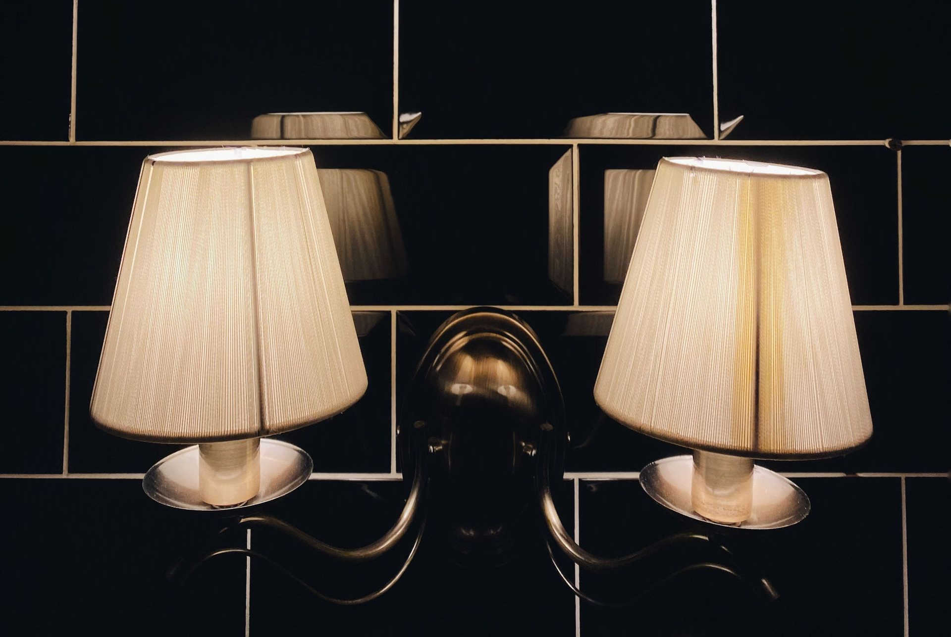 Two beige wall sconces with pleated shades on a dark tiled wall, glowing softly.