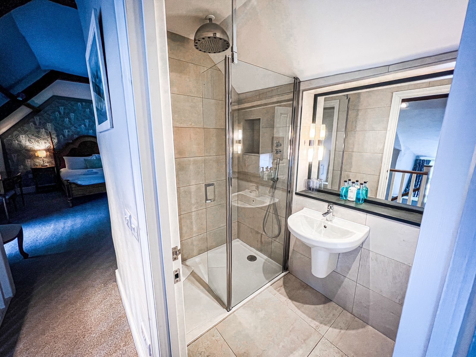 A modern bathroom with a glass-enclosed shower, pedestal sink, and large mirror, adjacent to a hotel bedroom.