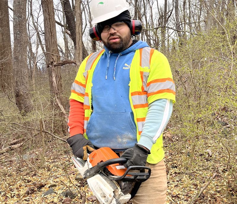 A man wearing a hard hat and safety vest is holding a chainsaw in the woods.