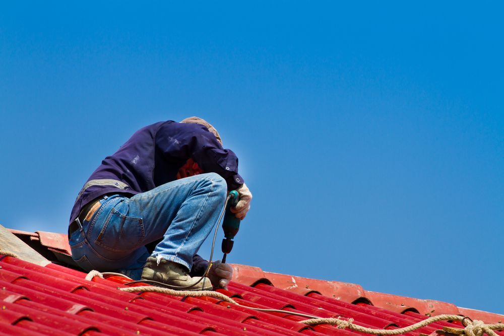 Roofer in Blue Jeans and Shirt, Using a Drill on A Red Roof Under a Clear Blue Sky — Geoff Hall Roofing in Kempsey, NSW