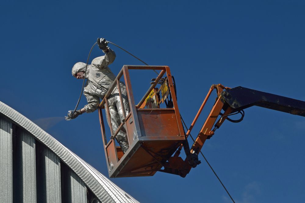 Person in Safety Suit Painting Building from A Boom Lift Against a Blue Sky — Geoff Hall Roofing in Kempsey, NSW