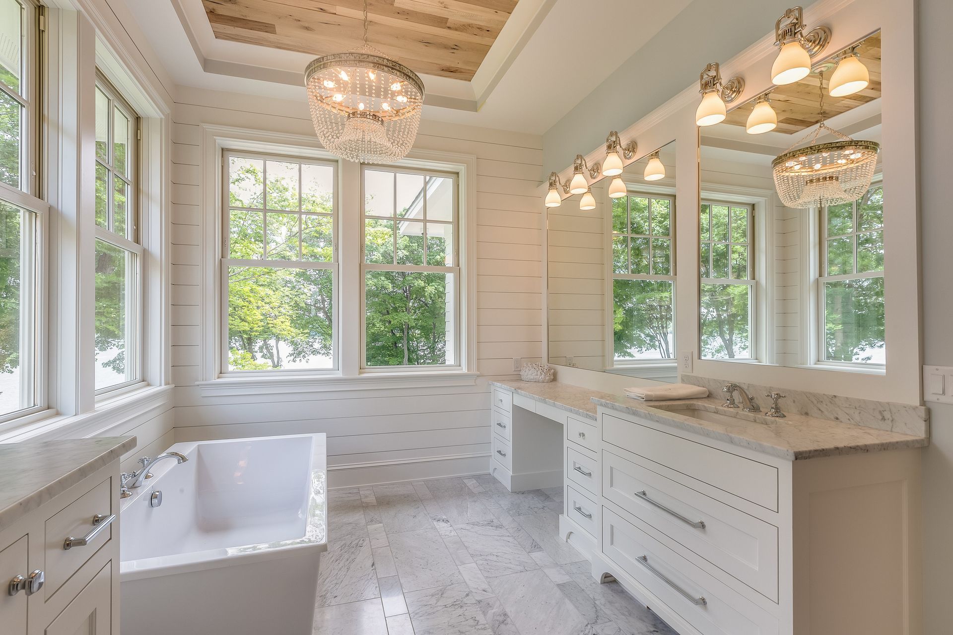 Elegant white bathroom with bathtub, vanity, and chandelier; light marble floor and wood ceiling.