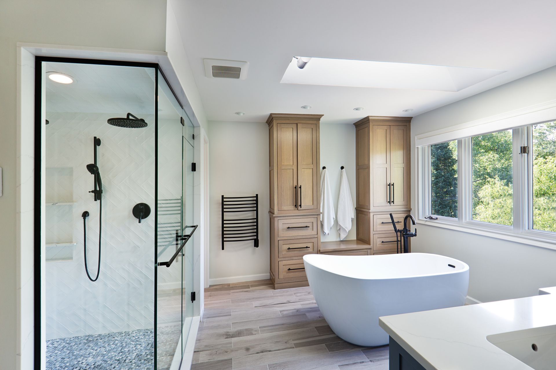 Modern bathroom with walk-in shower, soaking tub, and light wood cabinetry. Large window and skylight provide natural light.