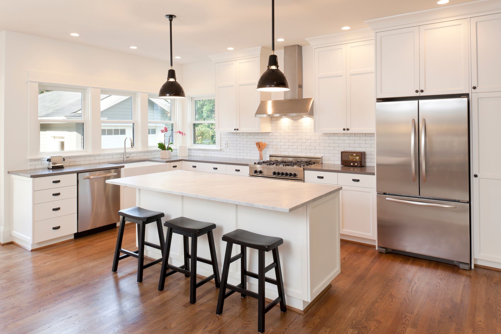 White kitchen with island, stainless steel appliances, black stools, and dark wood floors.