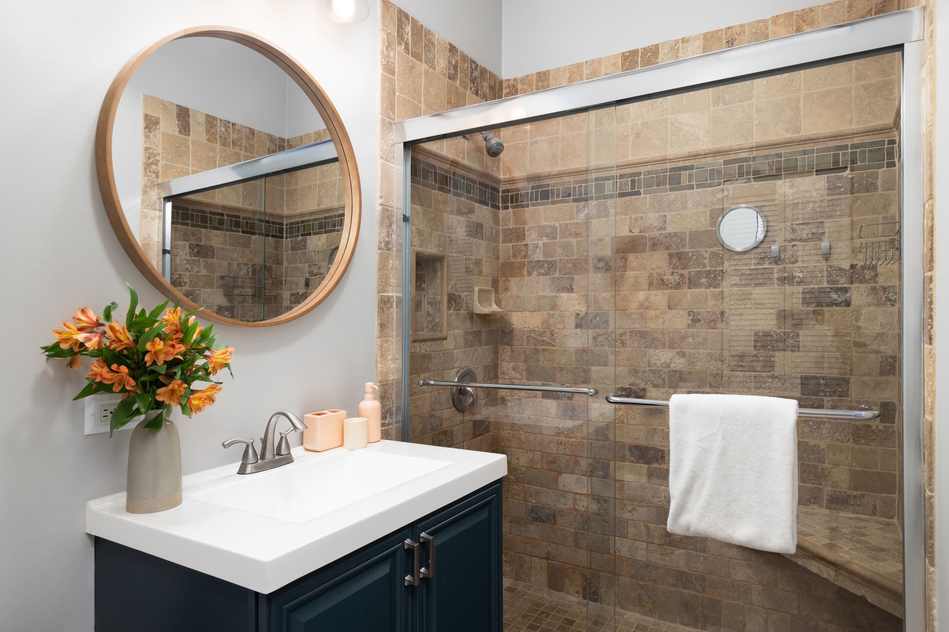 Bathroom with a vanity, a round mirror, and a tiled shower. Orange flowers and a white towel.