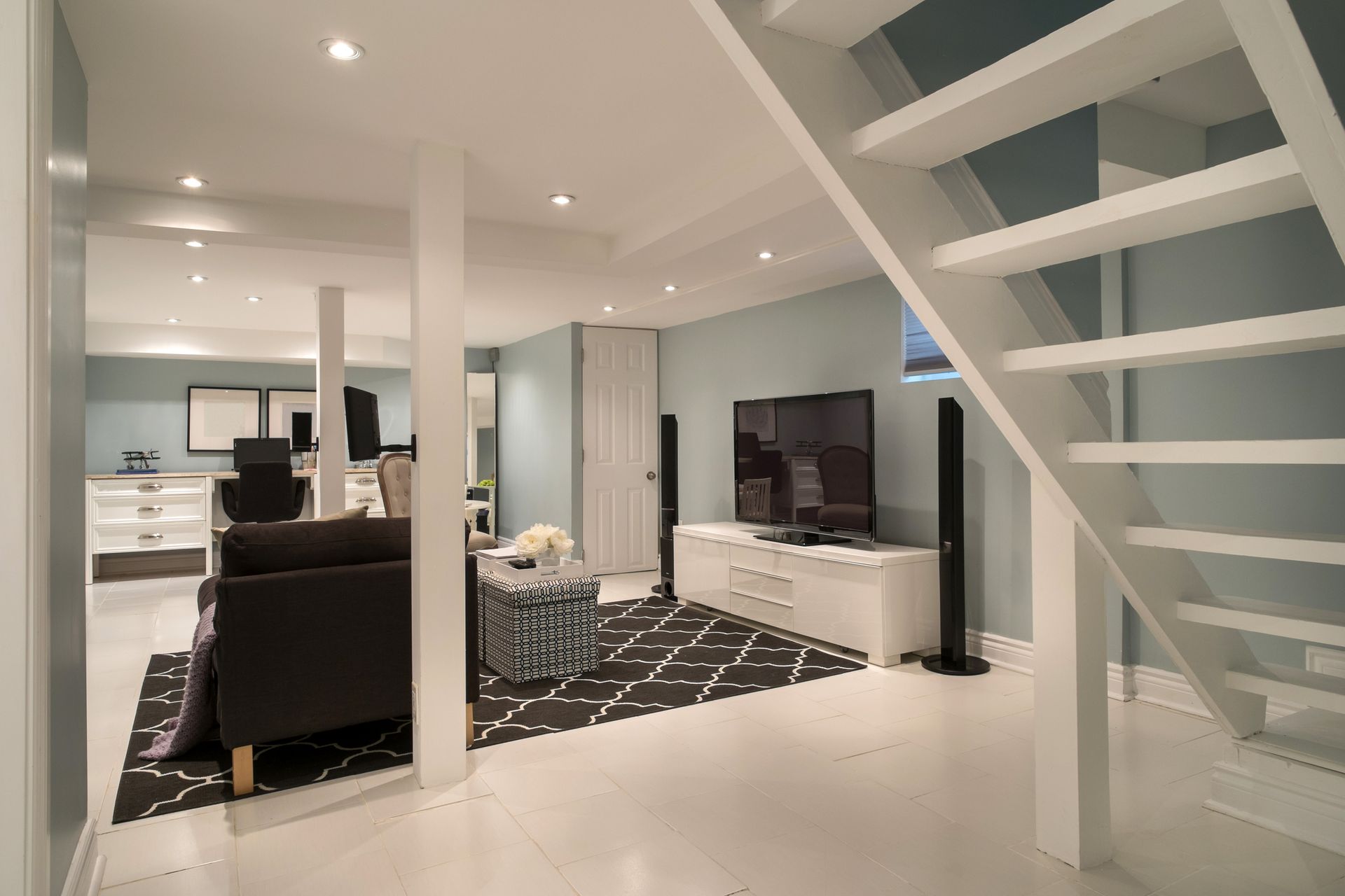 Basement living room with a couch, TV, and stairs. White walls, light blue accent wall, and a black and white rug.