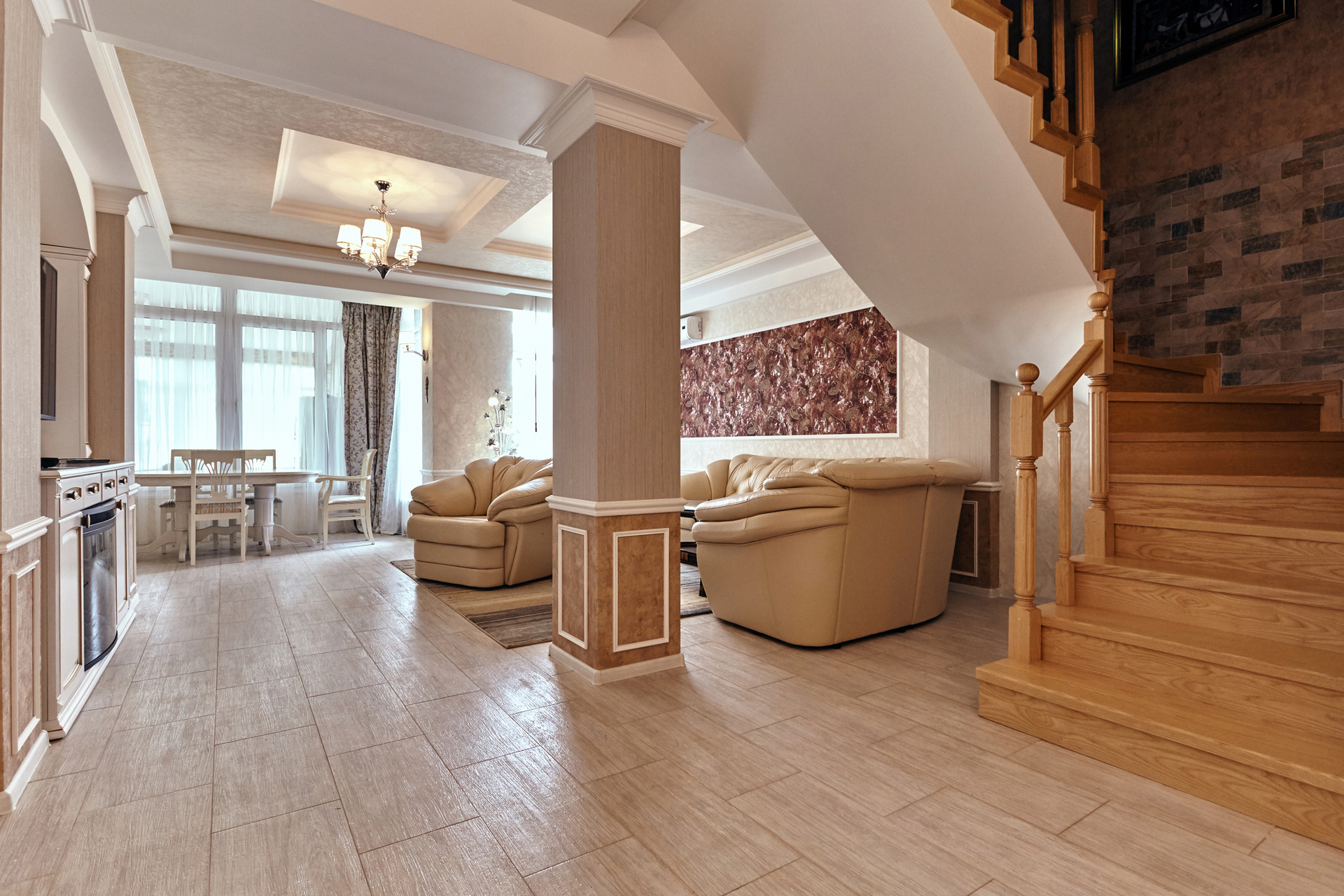 Spacious living area with light wood floors, staircase, beige couches, a dining area, and a central column.