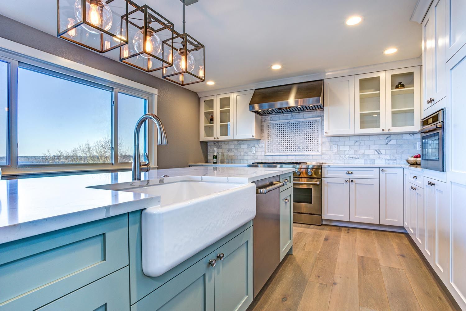 Modern kitchen with white and teal cabinets, marble countertops, and large window.