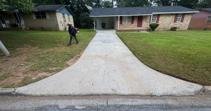 A person using a leaf blower on the grass beside a concrete driveway leading to a brick ranch-style house.