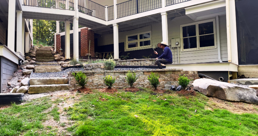 A person works on a stone retaining wall and garden bed alongside the steps of a tiered house deck.