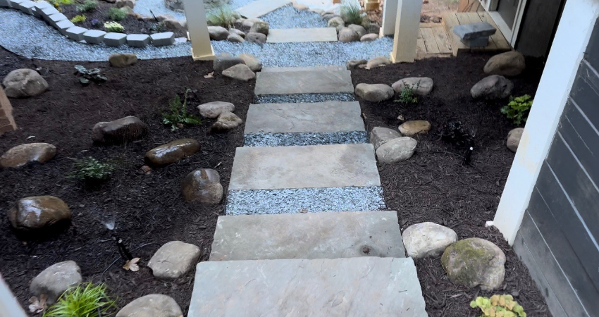 Outdoor stairs made of large stone slabs separated by gravel strips, surrounded by dark mulch and decorative rocks.