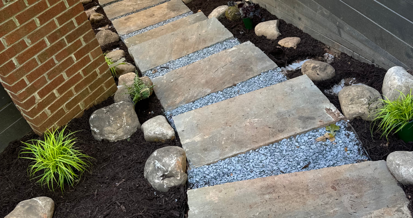 A garden path of rectangular stone pavers set in gray gravel, surrounded by dark mulch, decorative rocks, and plants.