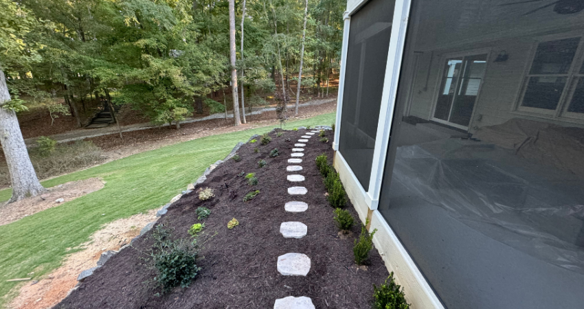 A stone pathway winds through a mulched garden bed beside the screened-in porch of a house near a grassy yard.