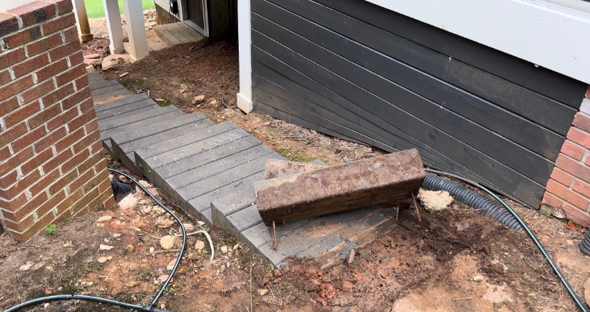 A wooden ramp leading to a house entrance, obstructed by a large, fallen timber beam on unfinished dirt ground.