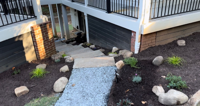 A stone pathway leads down a landscaped yard toward concrete steps beneath a raised wooden deck with a black railing.