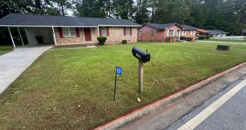 A single-story brick house with a carport and a manicured green lawn, viewed from the street with a mailbox in front.