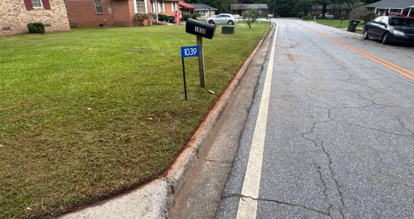 A residential street view showing a mailbox, a blue house number sign, a grassy lawn, and a road with a concrete gutter.