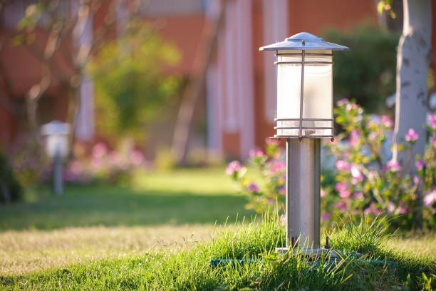 A modern outdoor lantern light stands in a grassy lawn with blurred pink flowers and a building in the background.