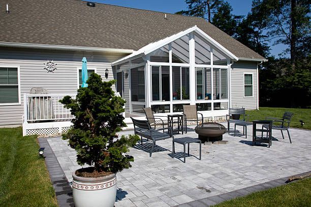 A backyard patio with light-colored pavers, a fire pit, several chairs, and a white sunroom attached to a gray house.