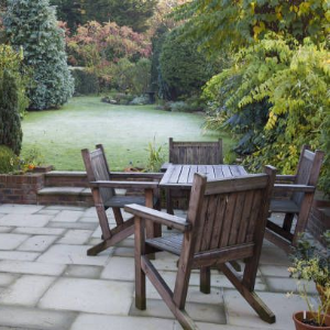 A patio with a wooden bench and dining set overlooking a grassy lawn surrounded by trees and shrubs.