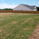 A large, fenced-in backyard with patches of green and brown grass under a bright blue sky.
