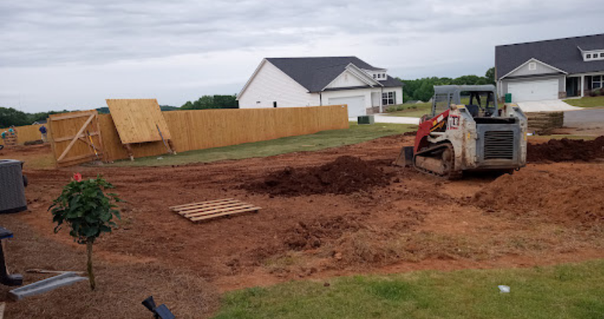 A skid steer loader sits on a dirt construction site in front of a wooden fence and houses under a cloudy sky.