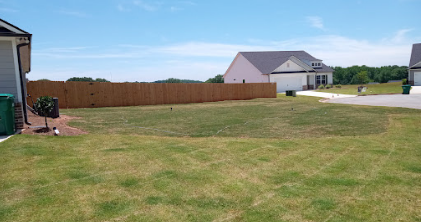 A suburban backyard with a wooden privacy fence, a patch of grass, and nearby houses under a clear blue sky.
