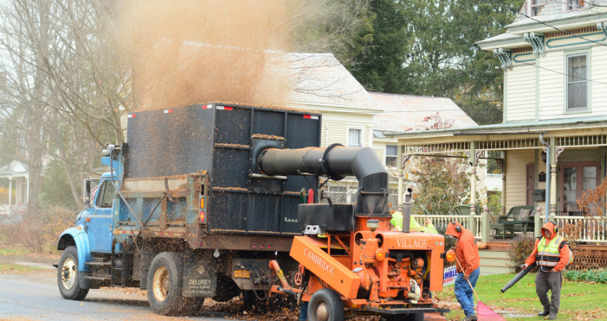 A blue truck and orange leaf vacuum clean up fallen leaves on a residential street with workers nearby.