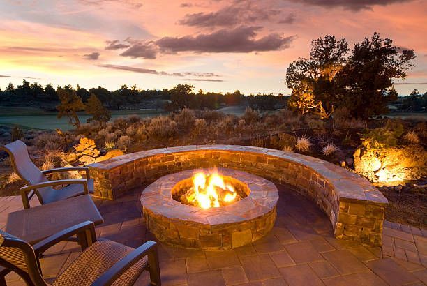 A lit stone fire pit with curved seating on a patio at sunset, overlooking a scenic, hilly landscape.