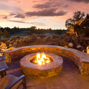 A stone fire pit with a glowing fire, surrounded by a curved stone bench and chairs, overlooking a landscape at sunset.