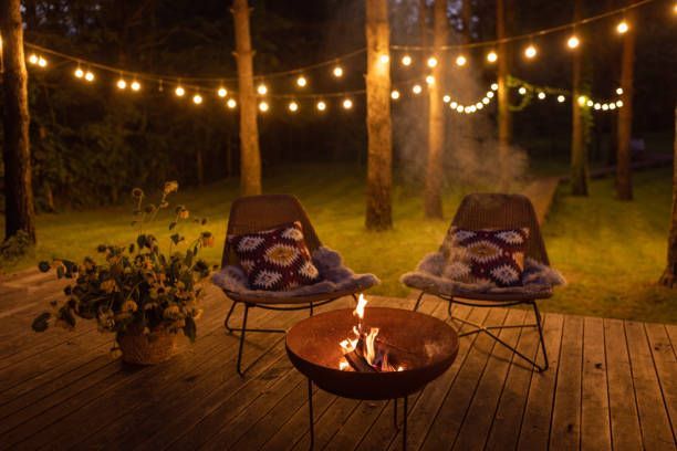 Two chairs with patterned pillows face a fire pit on a wooden deck under string lights in a forest at night.