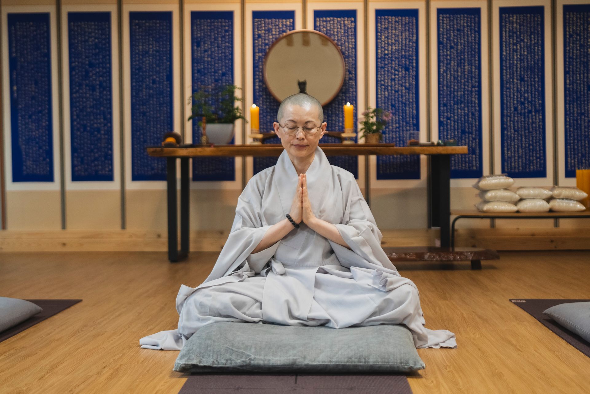 Woman in meditation pose, light clothing, seated on gray cushion, wooden floor. Interior, blue-paneled wall, altar, candles.