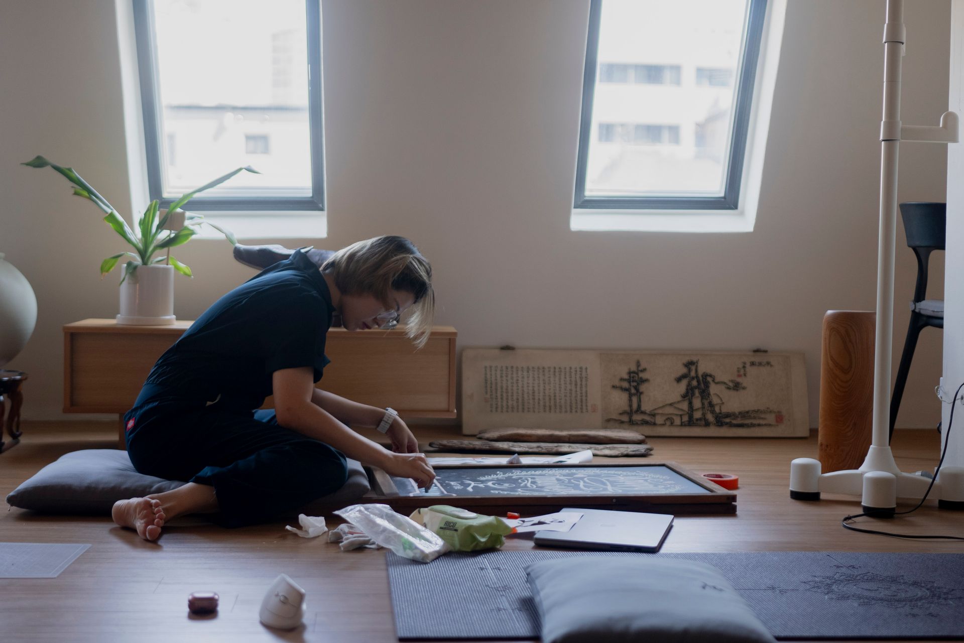 Person kneels on floor, working at a calligraphy board, near windows. Natural light fills the room.