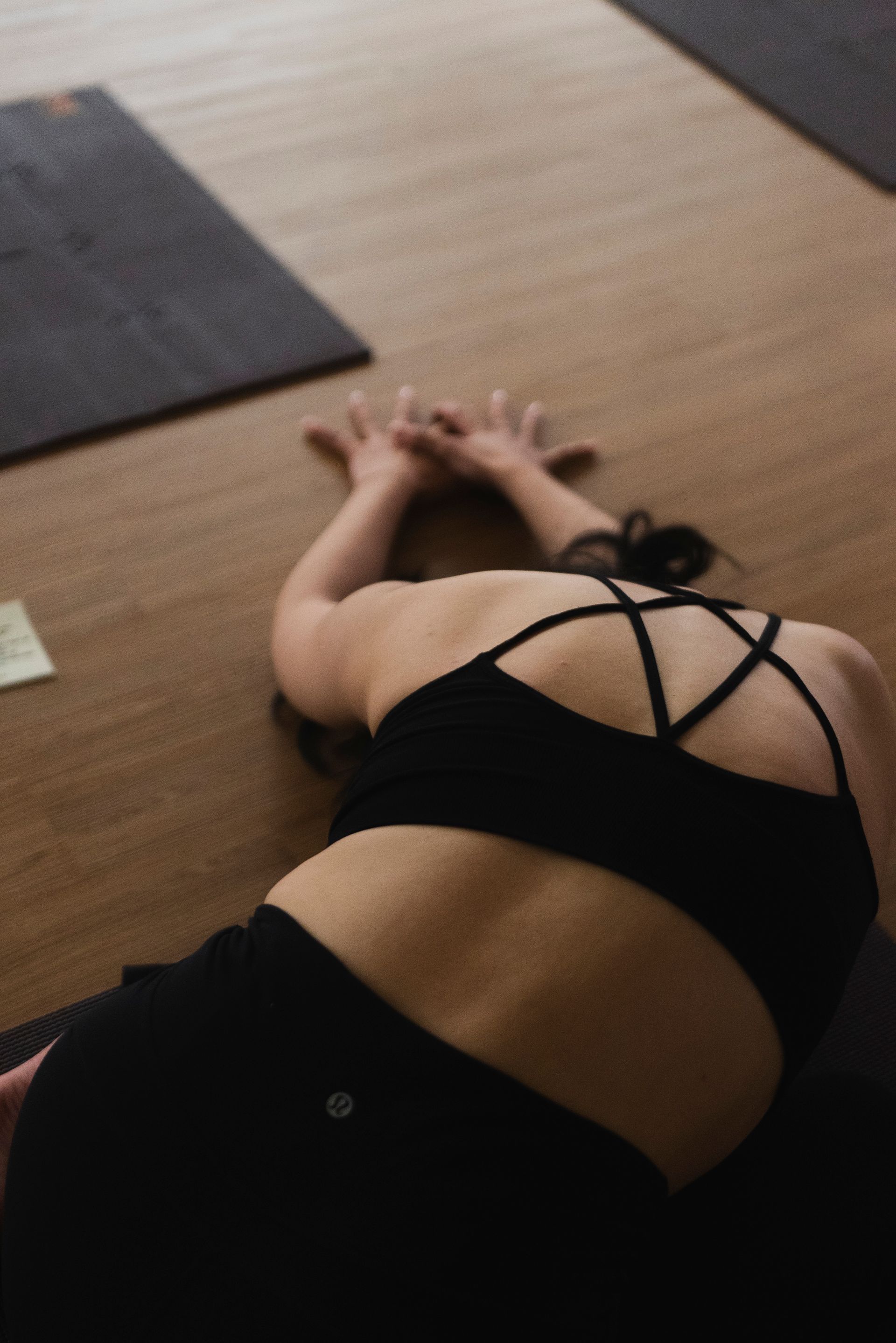 검은색 운동복을 입고 나무 바닥 매트 위에서 요가 자세를 취하고 있는 여성. 두 손을 머리 위로 모으고 있다.