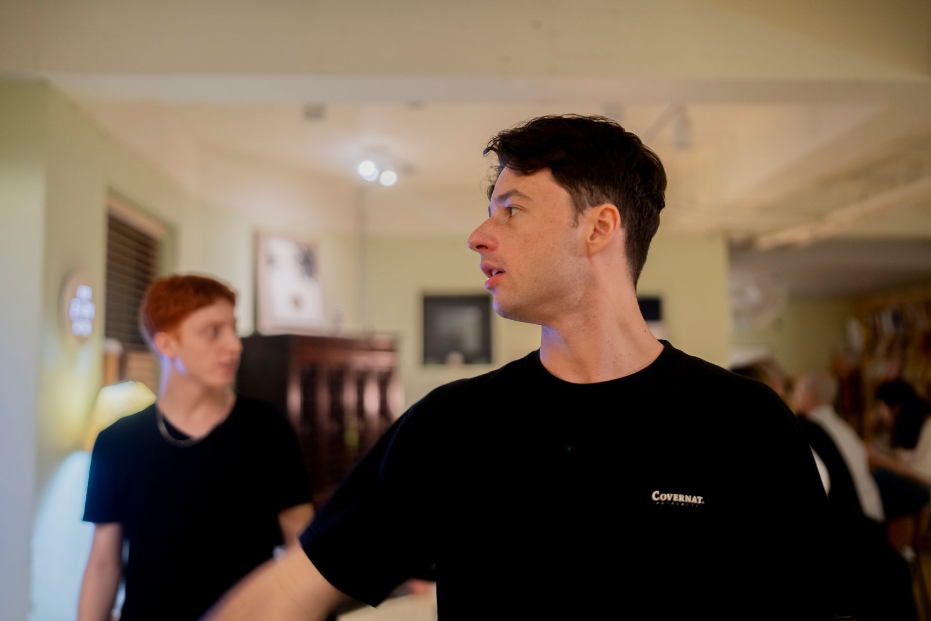 Two men in black shirts in a room; one gestures with arm extended, the other looks on.