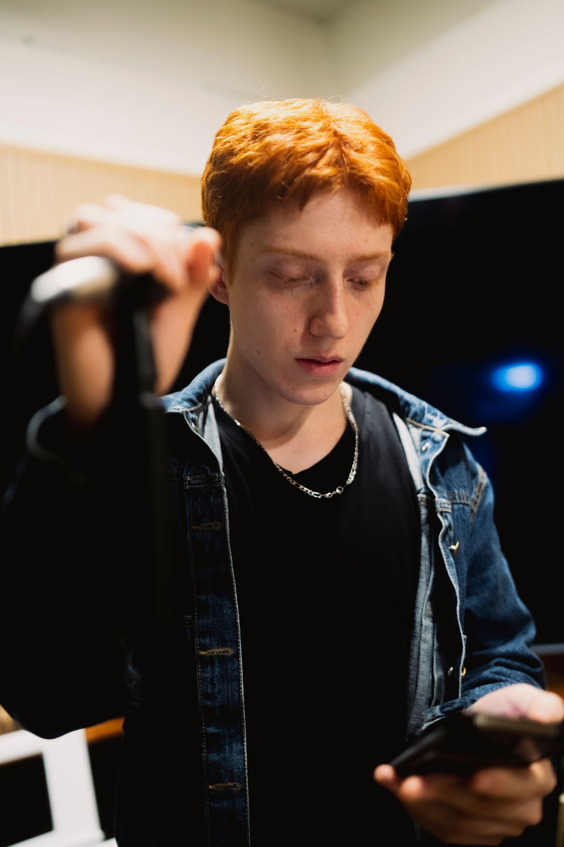 Young man with red hair, holding a microphone, looking at a phone, wearing a denim jacket.
