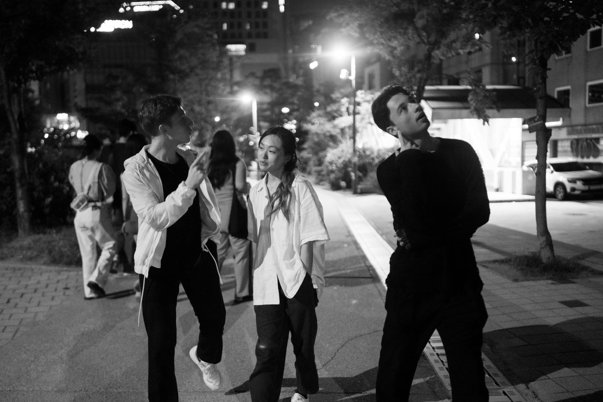 야경: 보도 위에 사람들이 모여 있다. 한 명은 위를 올려다보고, 다른 두 명은 앞으로 걸어가며, 가로등이 풍경을 비춘다.