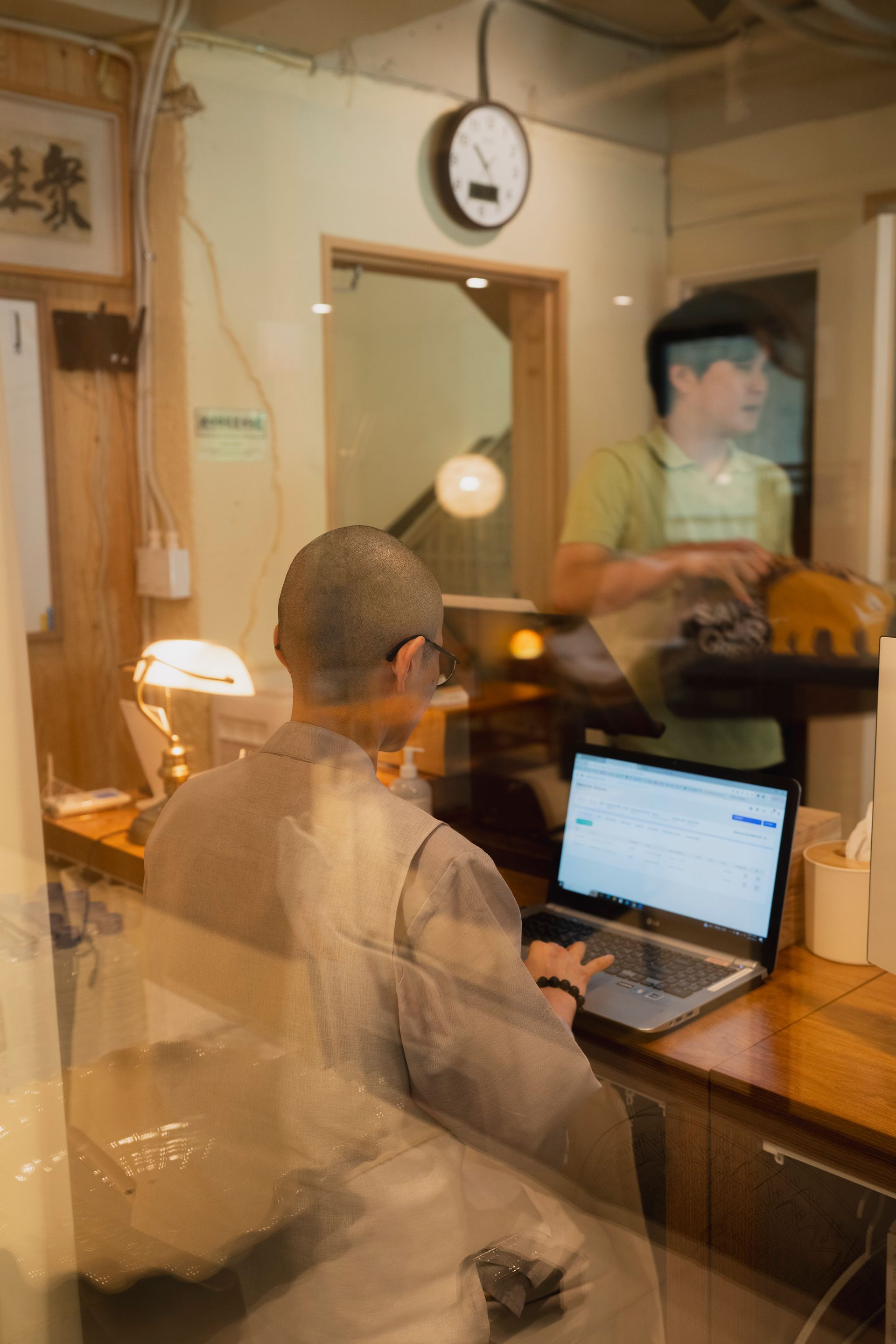 회색 가운을 입은 사람이 책상에서 노트북으로 작업하고 있습니다. 다른 사람이 뒤에서 걸어오고 있습니다. 실내입니다.