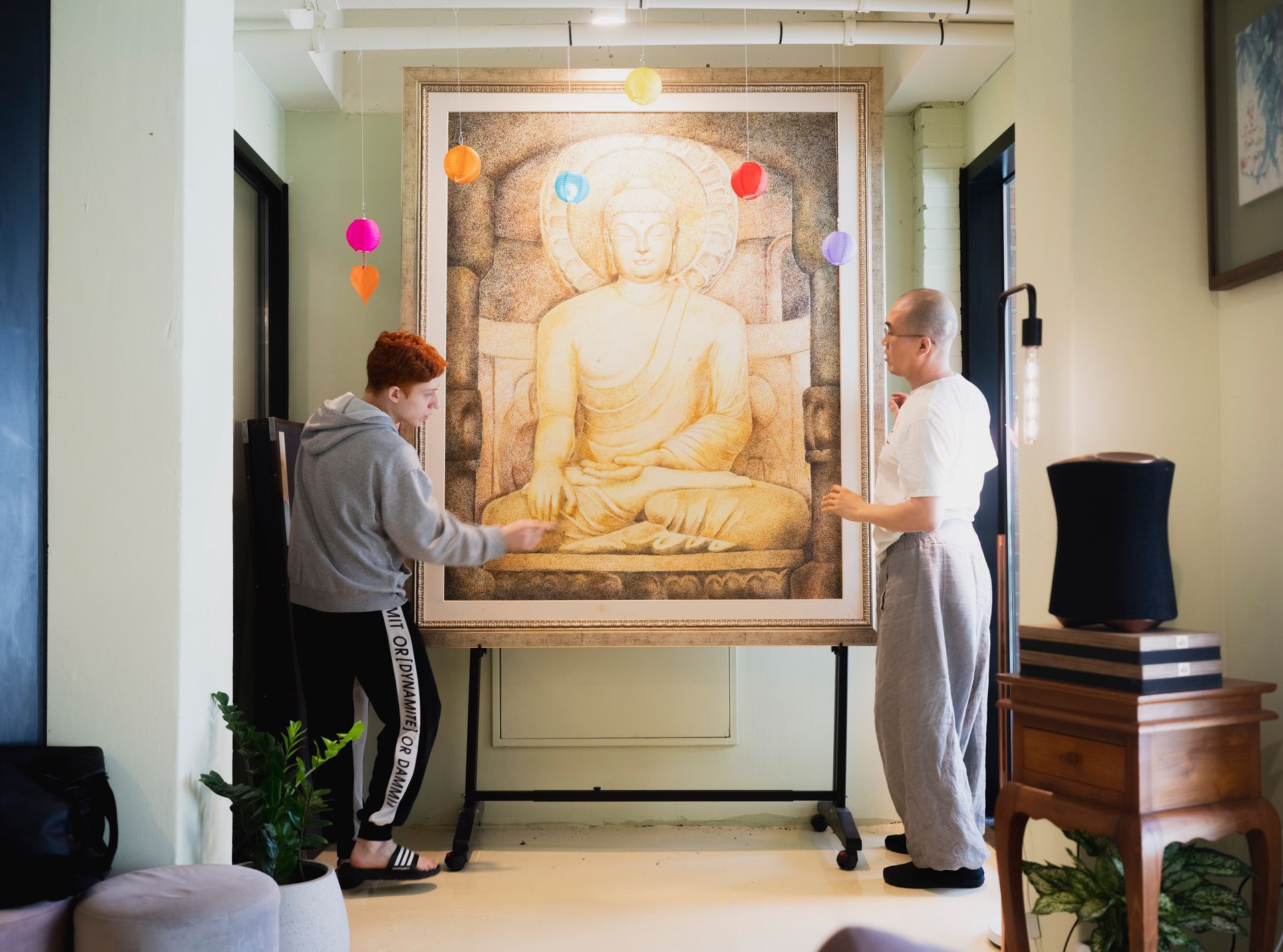 Two people hanging a large Buddha artwork in a room with colorful decorations.
