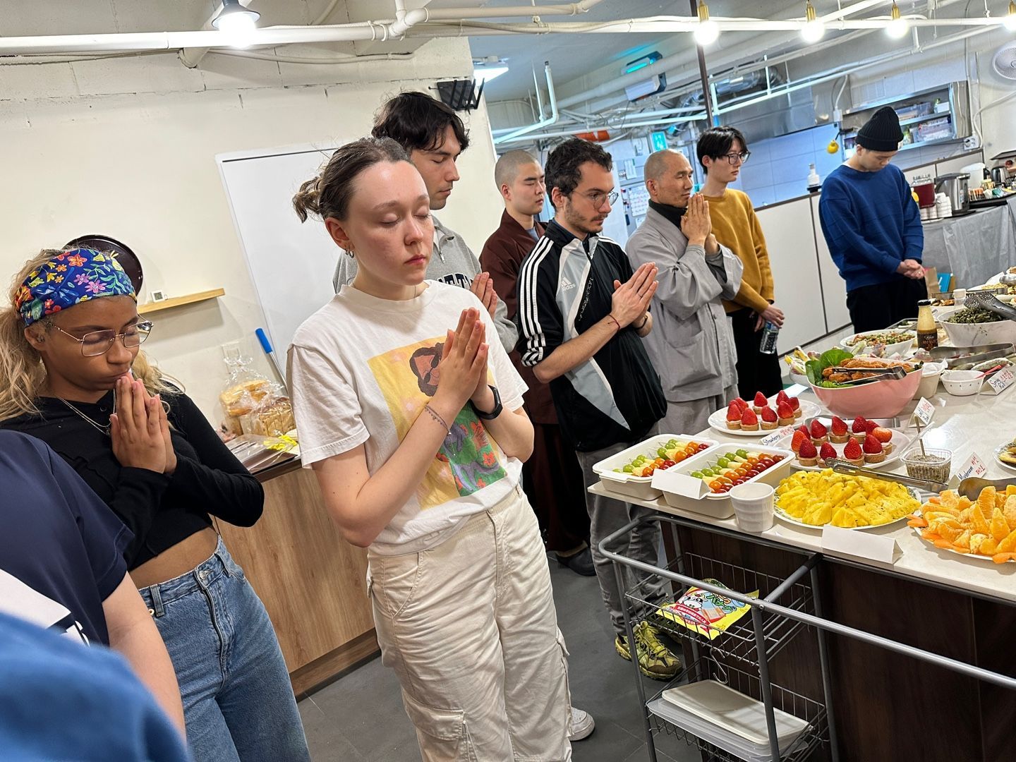 사람들이 음식이 가득 담긴 테이블 앞에서 손을 모으고 기도하는 모습. 실내는 환하게 불이 켜져 있다.