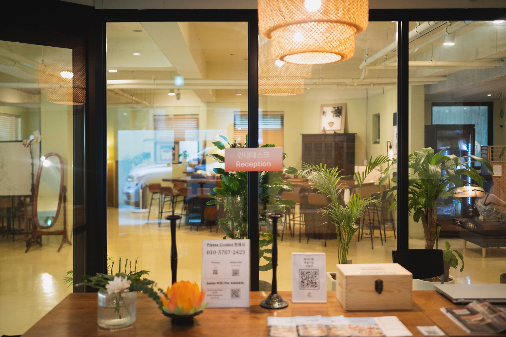 카페 입구. 나무 테이블과 장식, 싱그러운 식물, QR 코드 표지판. 좌석과 조명이 보이는 실내.