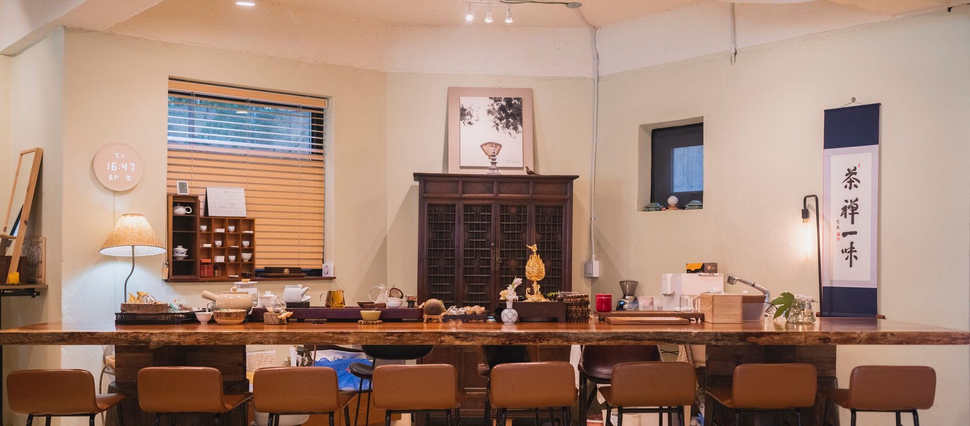 A tea room with a long wooden counter and stools, antique furniture, and a hanging scroll.