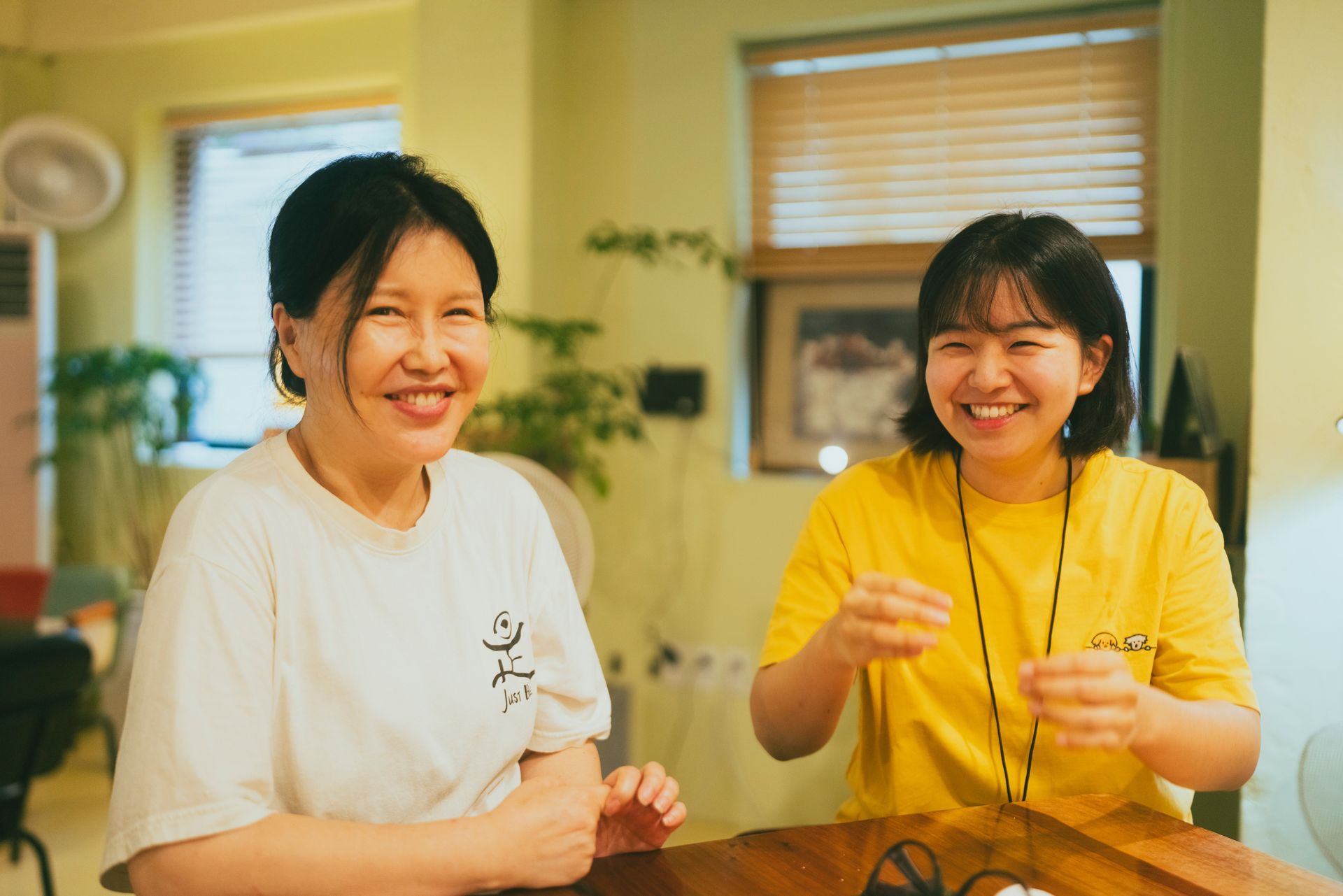 Two women smiling indoors, one in white shirt, one in yellow shirt, next to each other.
