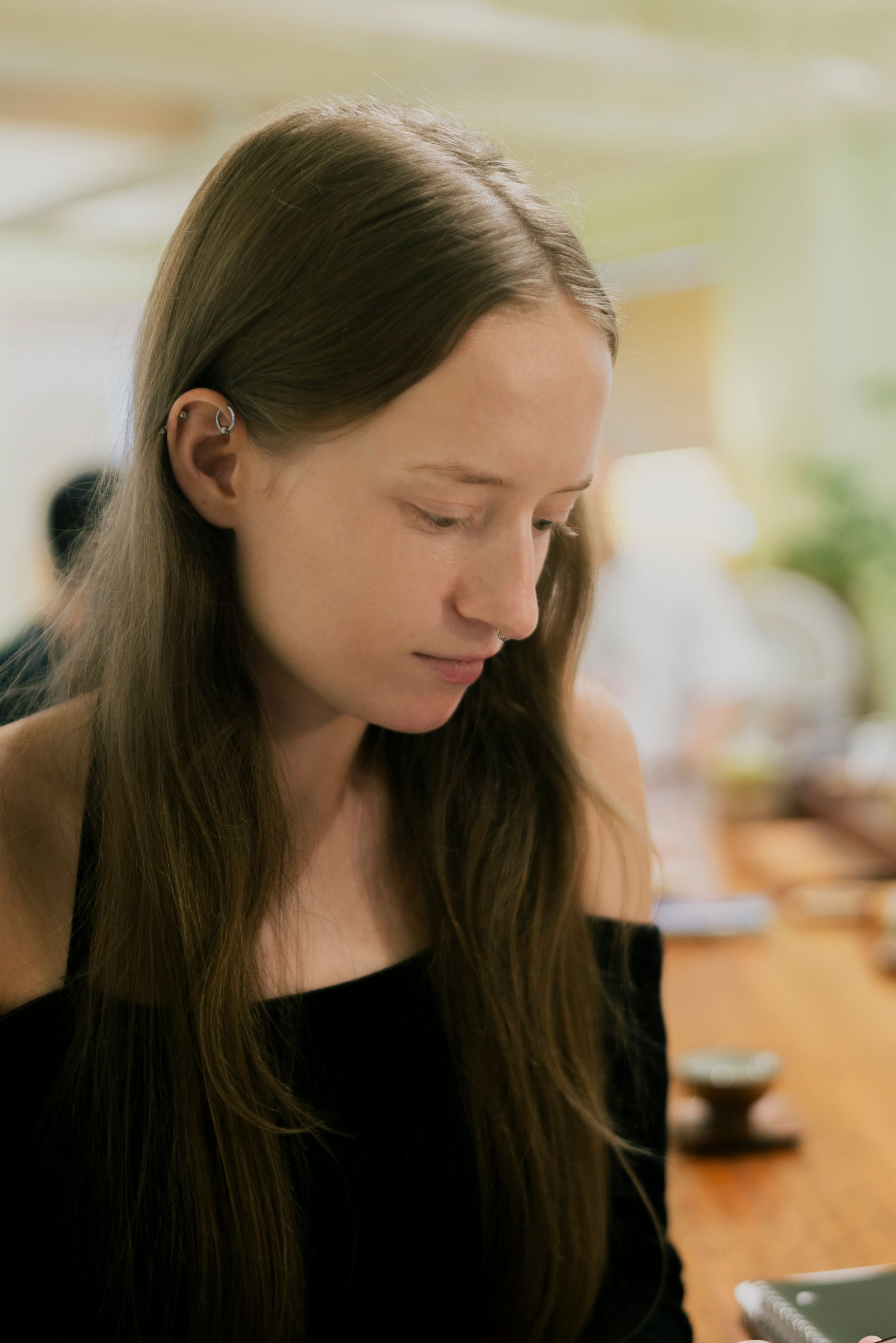 Woman with long brown hair, looking down, wearing black off-shoulder top; seated at a table.