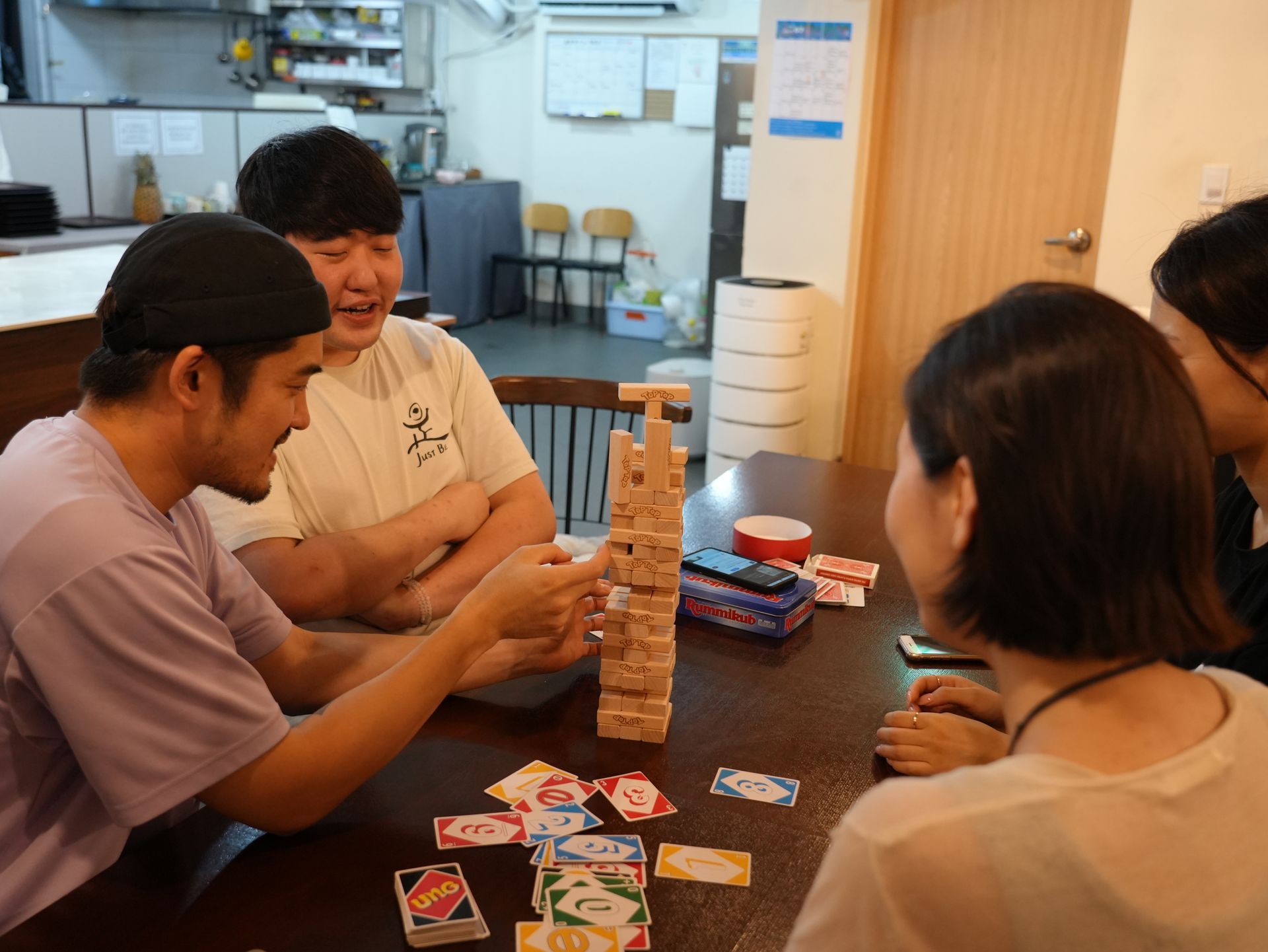 People playing Jenga and Uno at a table; a tower of wooden blocks stands tall as a player carefully pulls a block.
