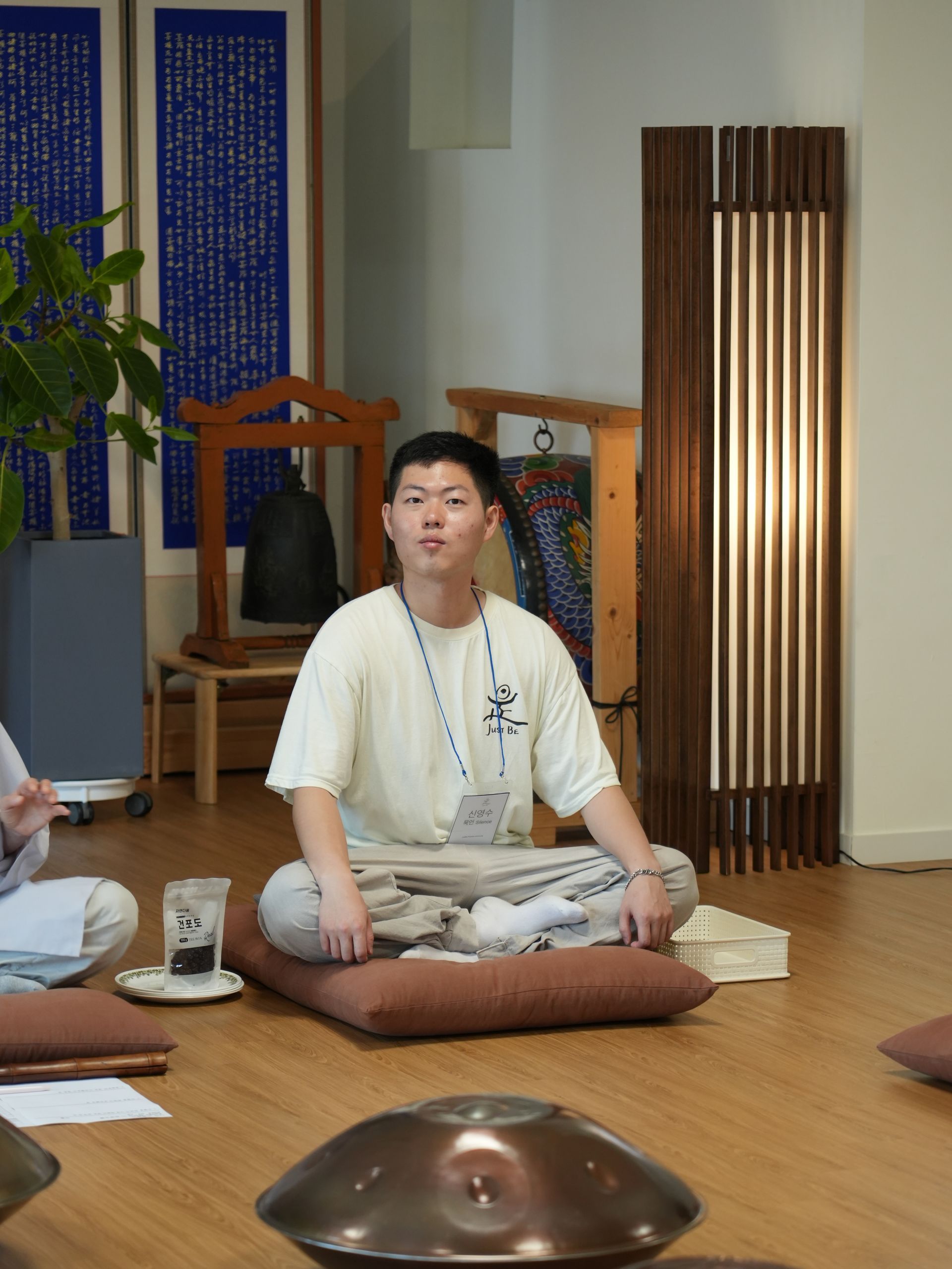 Man in meditation pose, seated on a cushion indoors, with a hang drum in front of him.