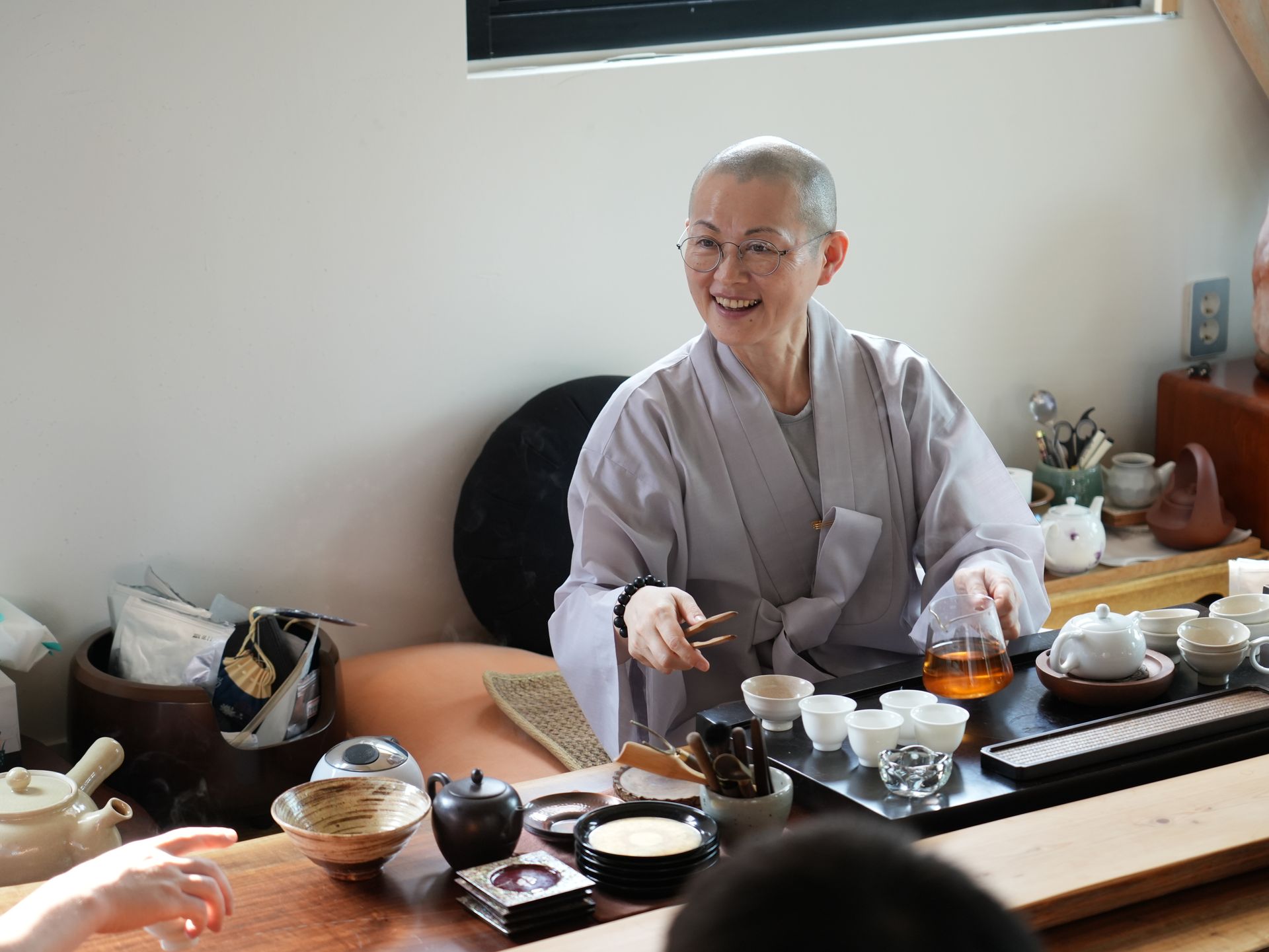 Smiling woman in gray robes, with a shaved head, prepares tea. Tea set and other items are on a table.