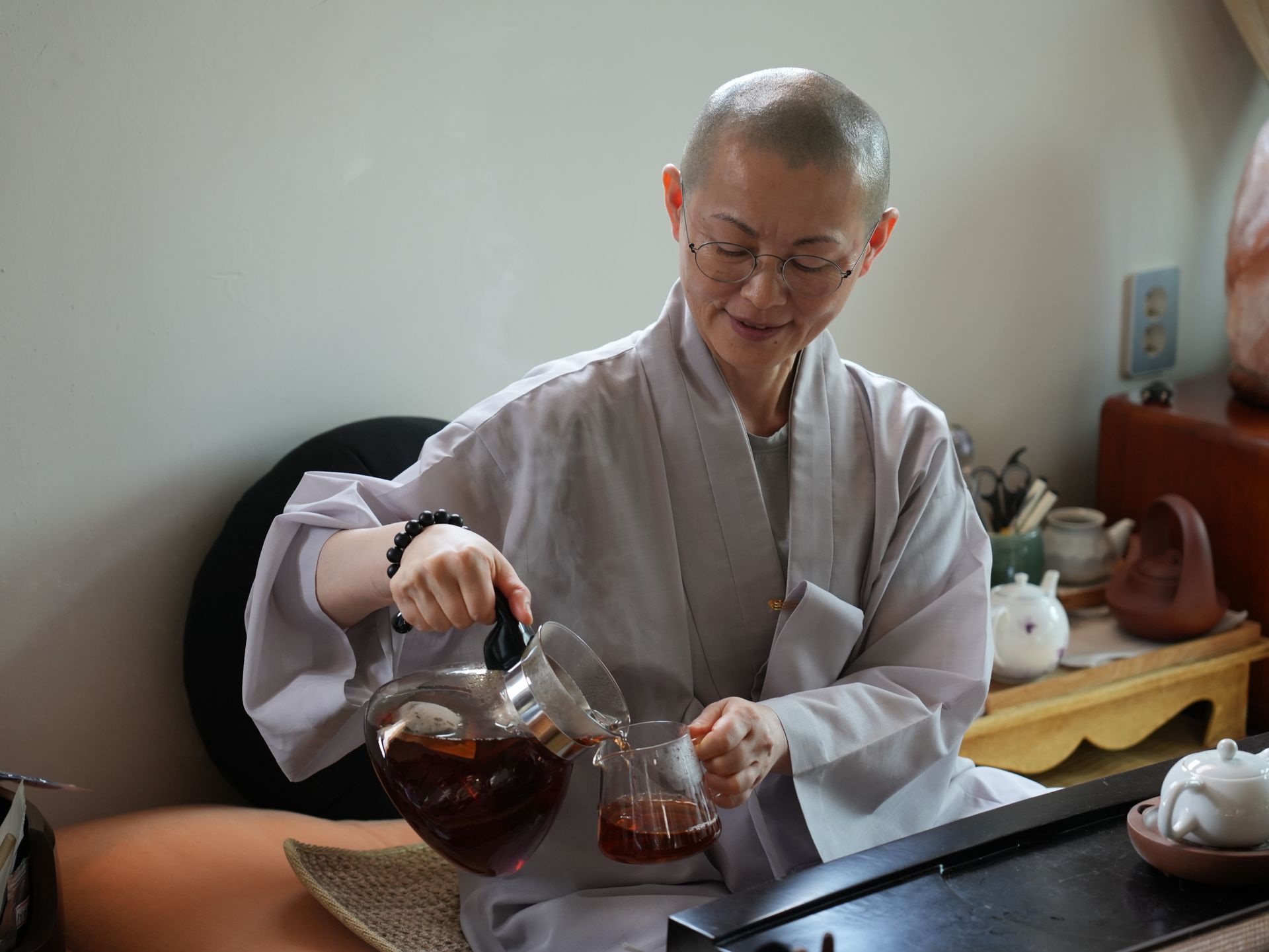 A bald woman wearing glasses and robes pours tea into a glass cup.
