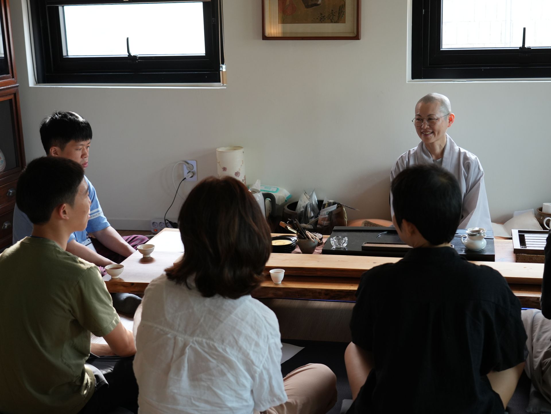 낮은 탁자 주위에 앉아 가운을 입은 미소 띤 사람의 말에 귀 기울이는 사람들. 자연광이 방 안을 가득 채운다.